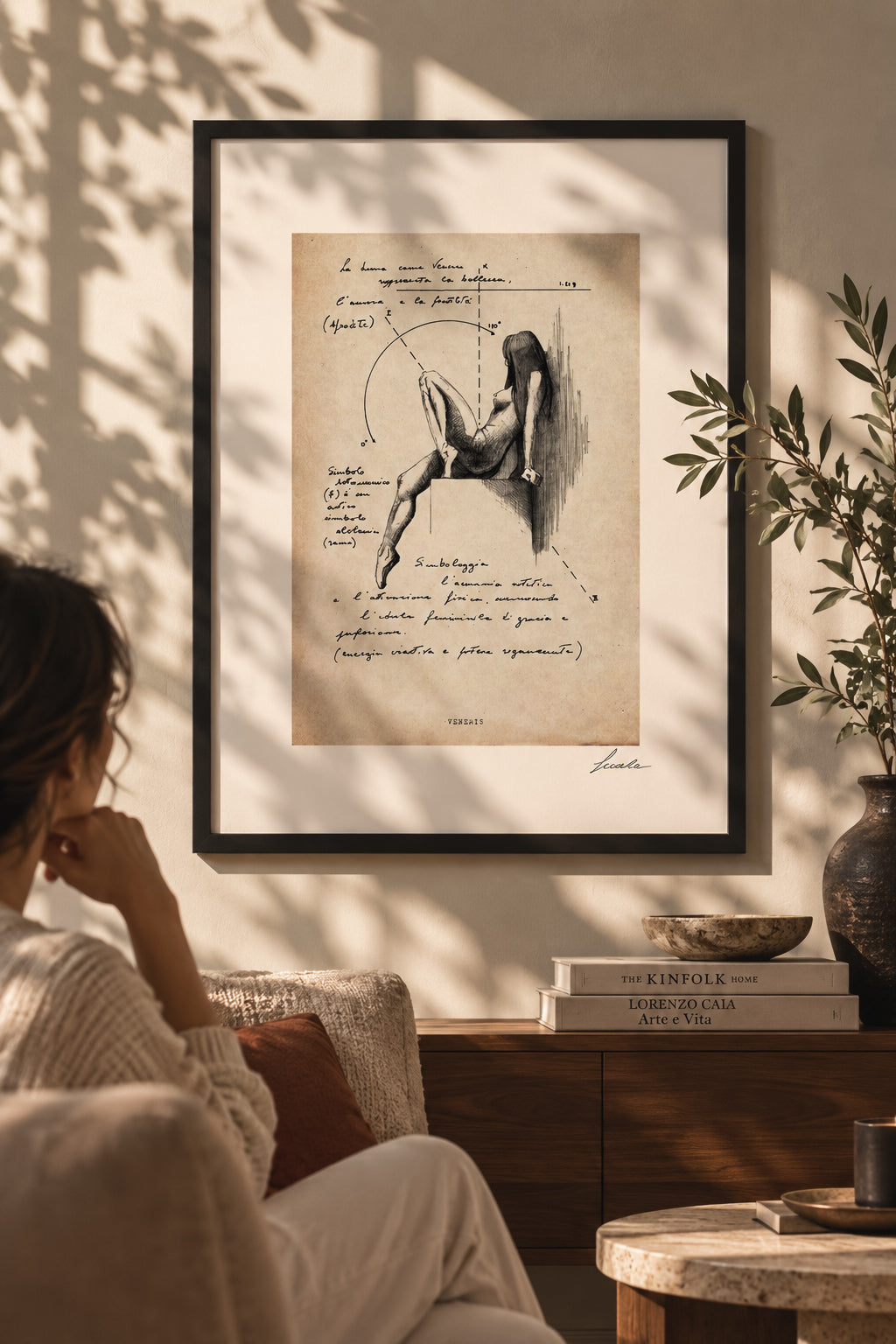 Veneris Archive ink print in a black frame on a sunlit wall, with a seated woman, books and plants in the foreground.