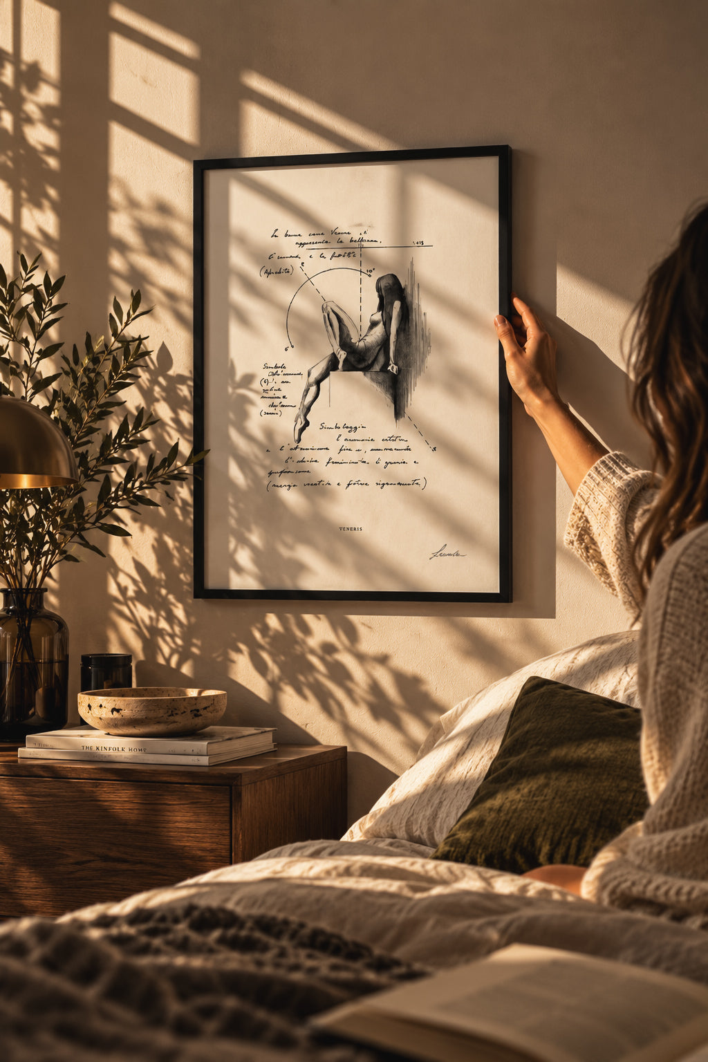 Woman holding a black-framed Veneris Studio ink print against a bedroom wall, illuminated by warm afternoon light.